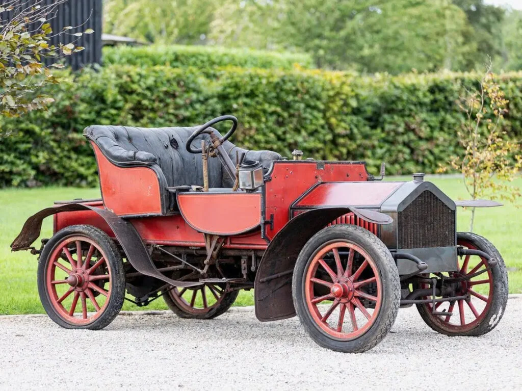 Unrestored red veteran two-seater with a black buttoned leather seat and red wooden-spoked wheels, parked on gravel in front of a hedge