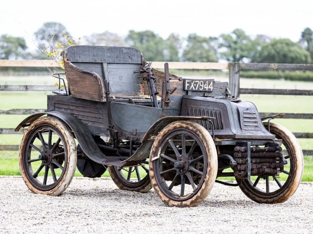 Unrestored circa-1903 Swift veteran car with faded dark paint, wooden-spoked wheels and the registration FX 7944, parked on gravel in front of a paddock fence