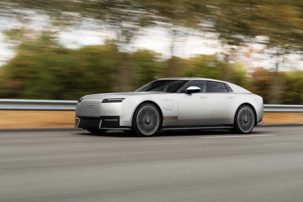 Silver Jaguar Type 01 electric GT driving on a test track, front three-quarter view, with a slatted front panel, slim headlights and a blurred background of trees