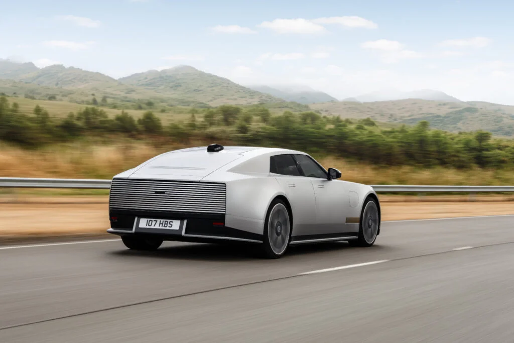 Silver Jaguar Type 01 driving on a test track with hills behind, rear three-quarter view, showing its slatted, windowless rear panel