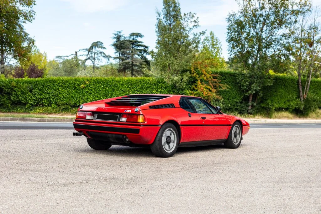 Rear three-quarter view of a red BMW M1 with louvred engine cover, parked on tarmac in front of a hedge and trees