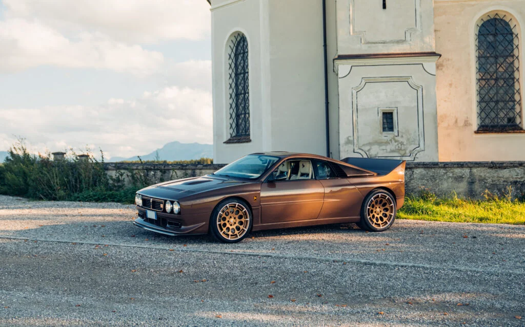 Brown Kimera EVO37 with gold wheels parked on gravel beside a white church with mountains in the distance