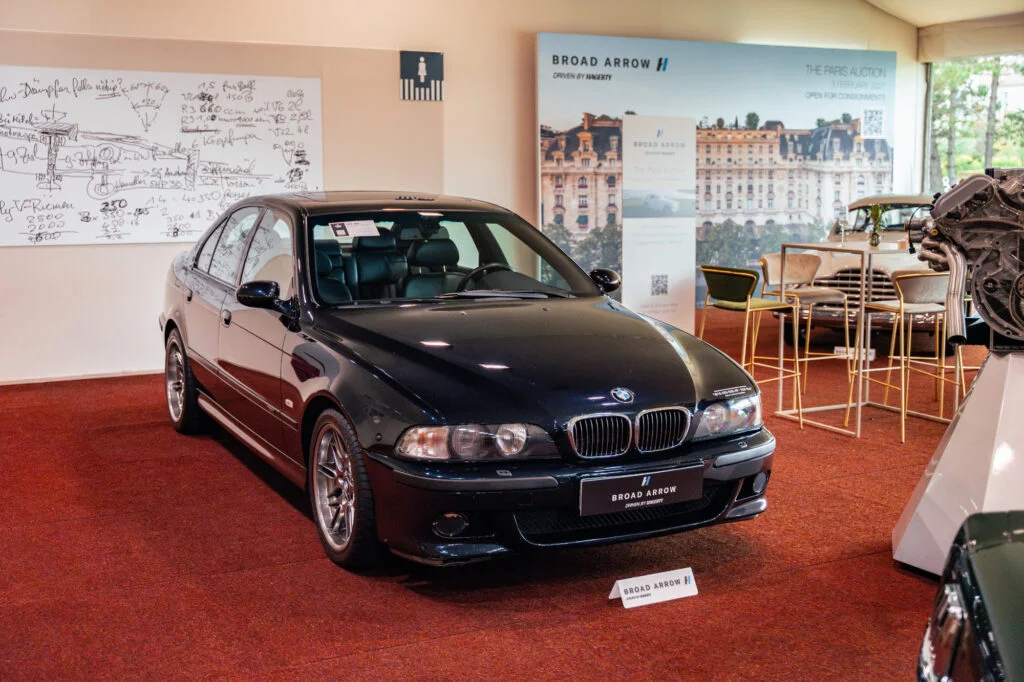 Black BMW M5 E39 on display at the Broad Arrow auction, beside a whiteboard covered in handwritten engineering notes and a spare engine on a stand