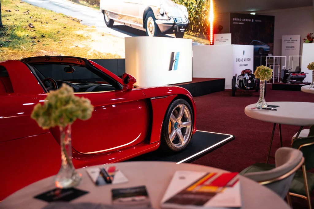 Red Porsche Carrera GT on a lit display plinth in the Broad Arrow auction marquee, seen past a table with catalogues and flowers