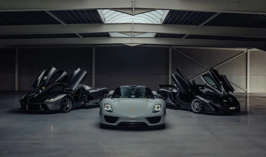 Grey Ferrari LaFerrari, grey Porsche 918 Spyder and dark grey McLaren P1 side by side in a hangar, the Ferrari and McLaren with their doors raised