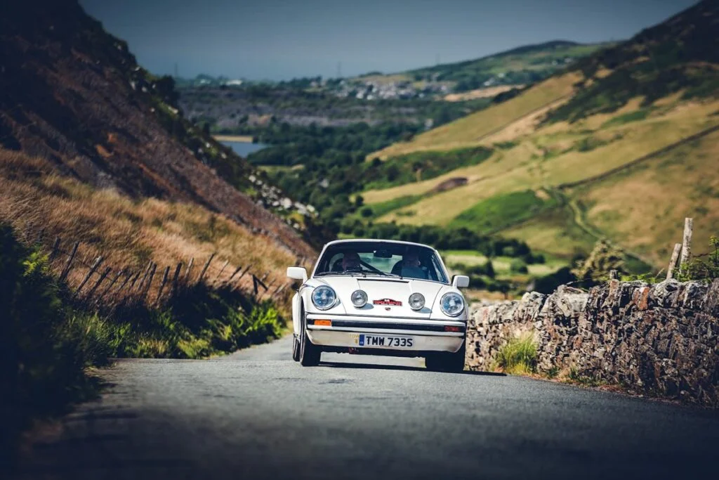 White Porsche 911 with rally plate driving along a narrow lane beside a dry-stone wall, with a valley, lake and village below