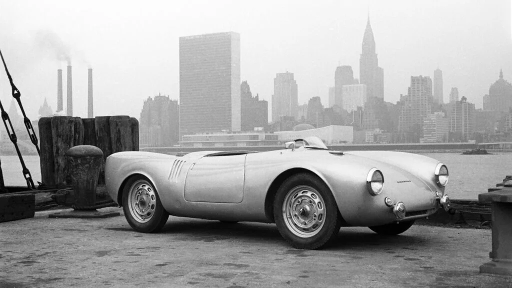 Black and white photograph of a silver Porsche 550 Spyder parked on a New York pier in 1953, with the Manhattan skyline and Chrysler Building behind