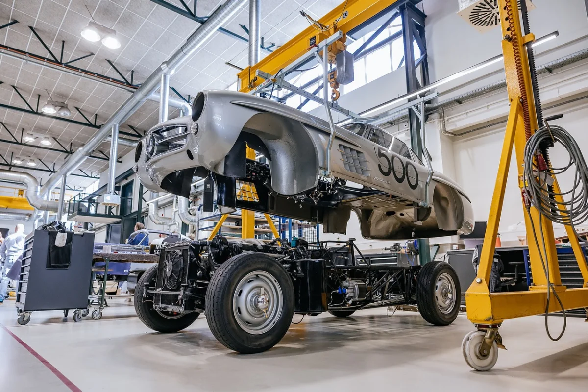 Silver Mercedes-Benz 300 SL body carrying race number 500 suspended from a yellow gantry crane above its bare chassis in the Classic Center workshop
