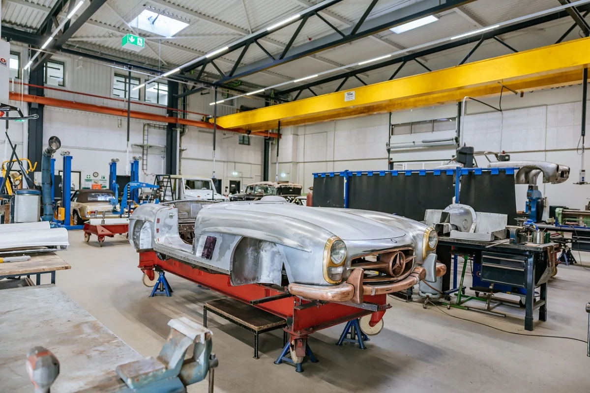 Bare aluminium-coloured Mercedes-Benz SL roadster bodyshell on a red trolley in the Classic Center bodyshop, with further cars under restoration behind